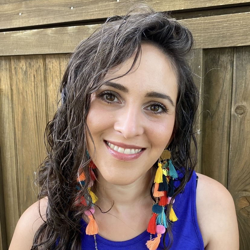 Woman with dark brown hair and some gray hairs showing, brown eyes, soft smile, light tan skin, colorful earrings and blue top.